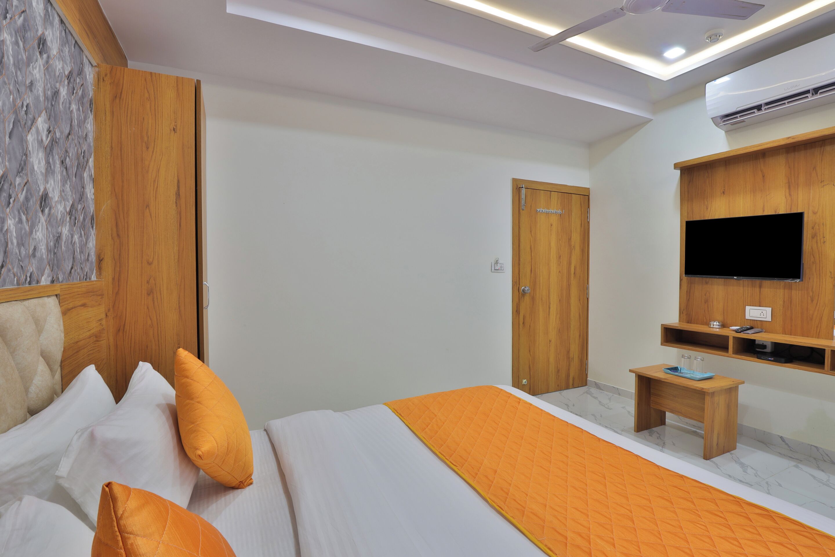 A modern hotel room with a bed, a wooden wardrobe, a small wooden side table, a television mounted on the wall, and an air conditioning unit.