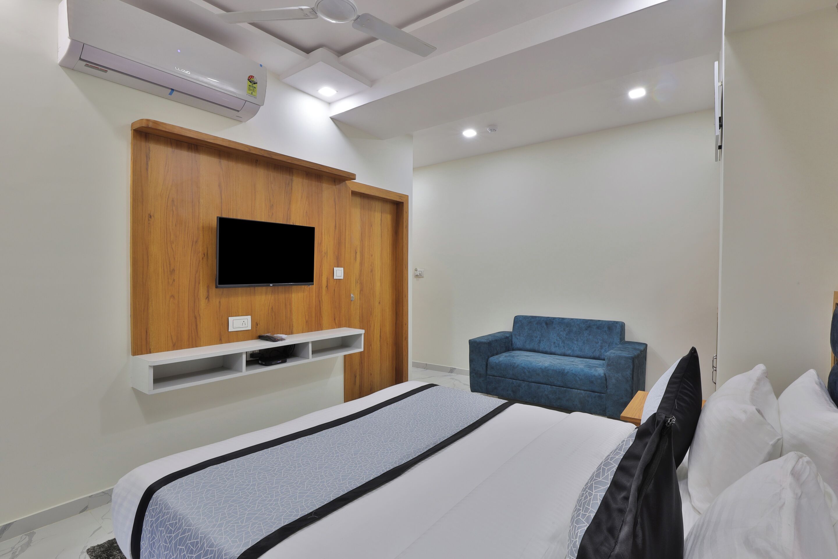 Ein modernes Hotelzimmer mit einem Bett, einem blauen Sofa, einem wandmontierten Fernseher und integrierten Regalen.