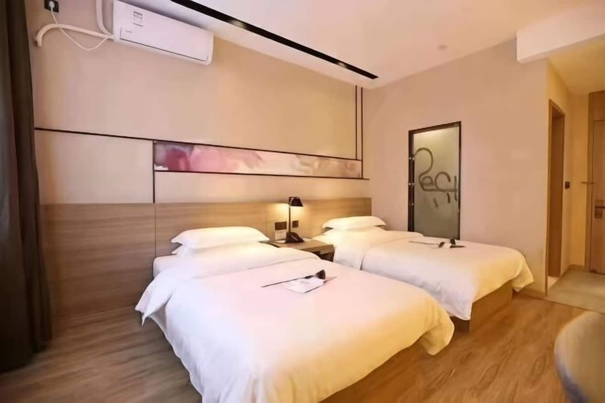 A hotel room with two beds, a bedside table with a lamp, a wall-mounted air conditioner, and a door leading to another room.