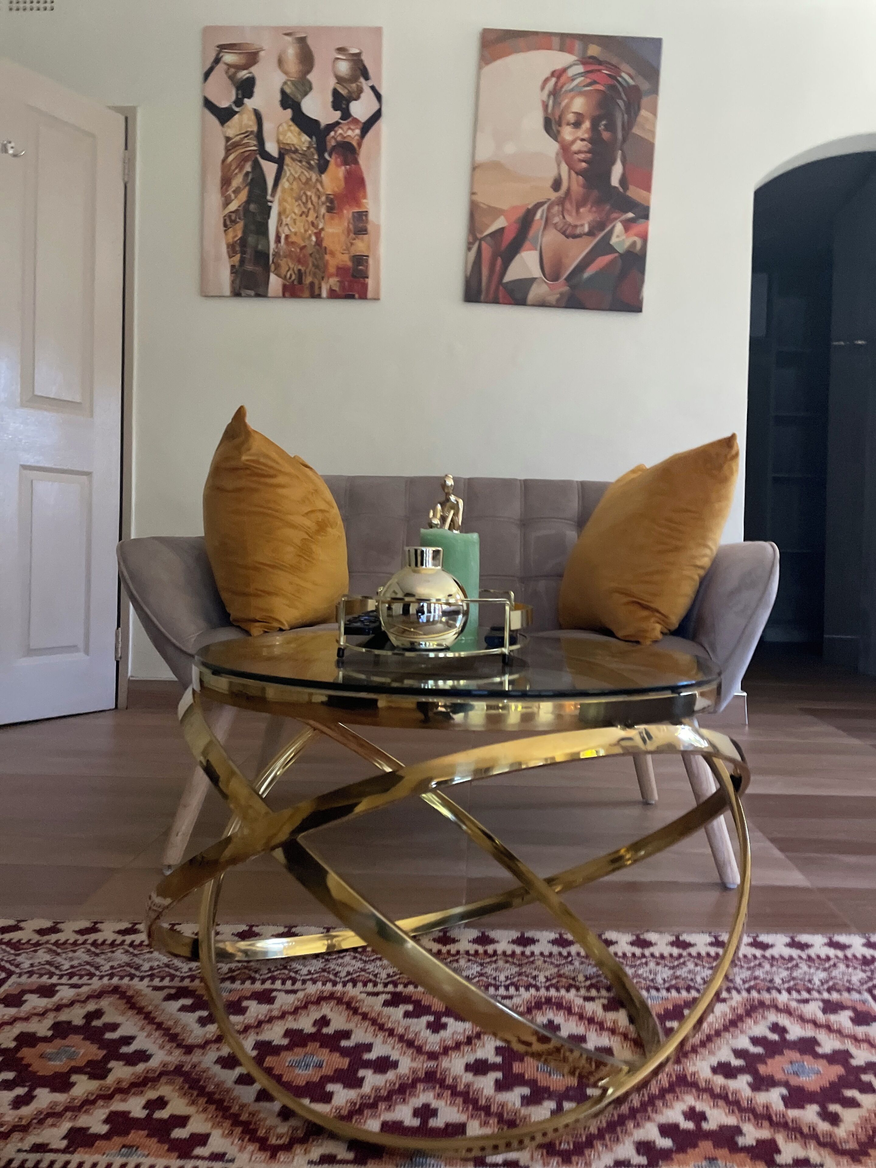 A living room with a glass coffee table, two gold-colored armchairs, a patterned rug, and two wall art pieces depicting cultural scenes.