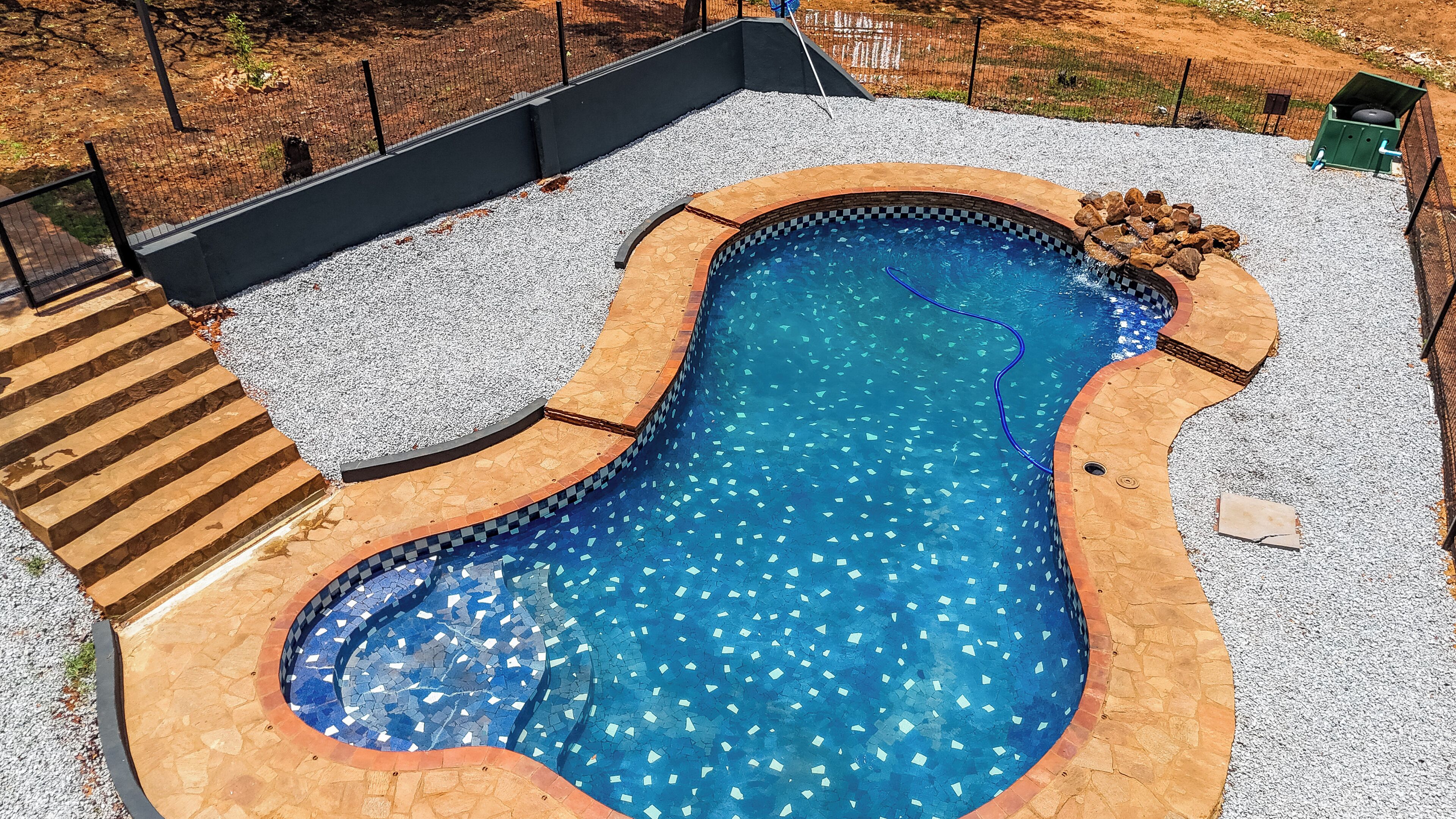An outdoor swimming pool with a unique shape, surrounded by a pebble border and a wooden deck with steps leading into the pool.