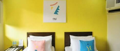 A hotel room with two beds, each with white bedding and a pillow featuring a logo. A small table with a water bottle and a menu is visible. The room has a yellow wall and a framed picture above the beds.