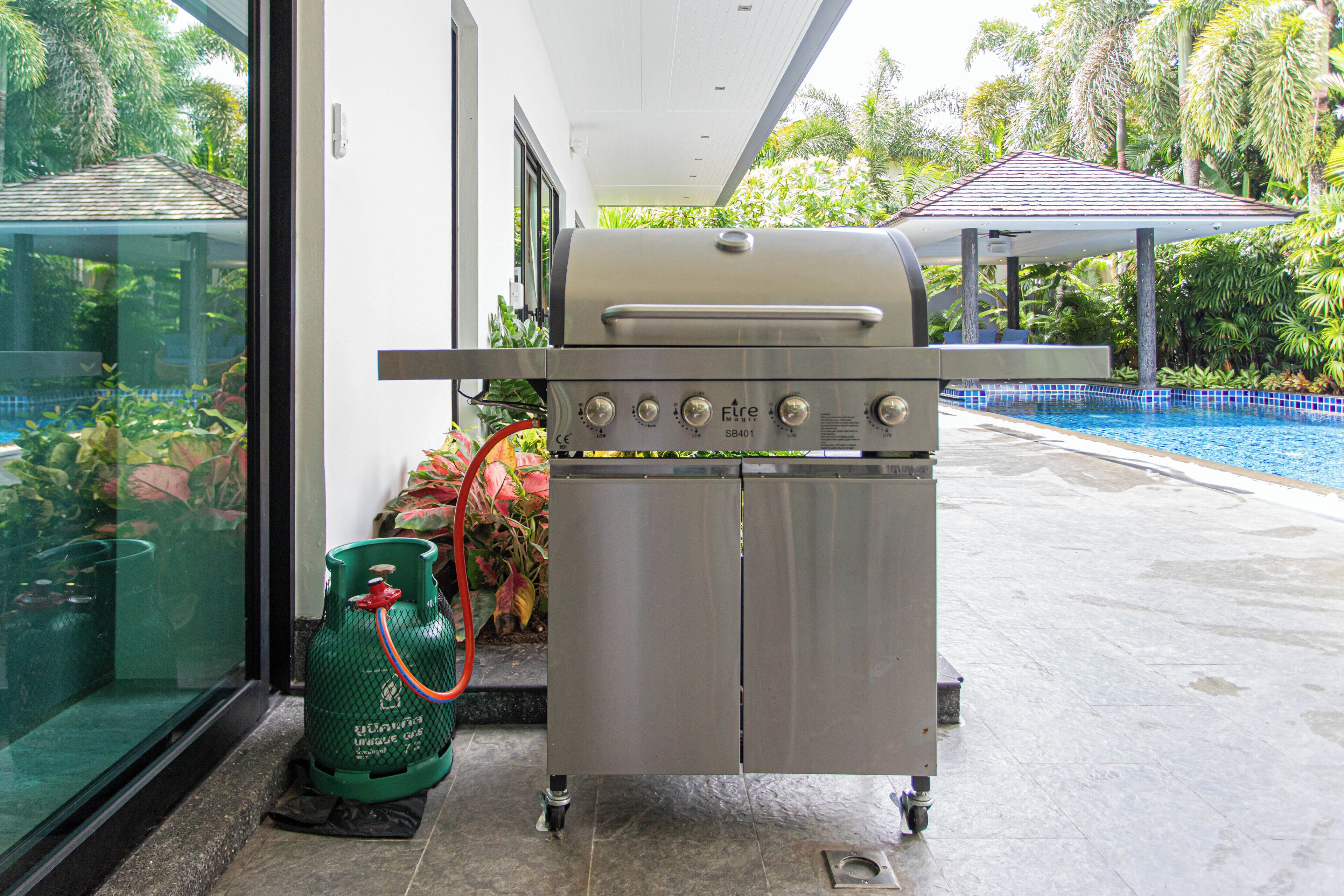 Un barbecue en acier inoxydable, équipé d’une bouteille de gaz et d’un tuyau, placé près d’une porte vitrée et d’une piscine.
