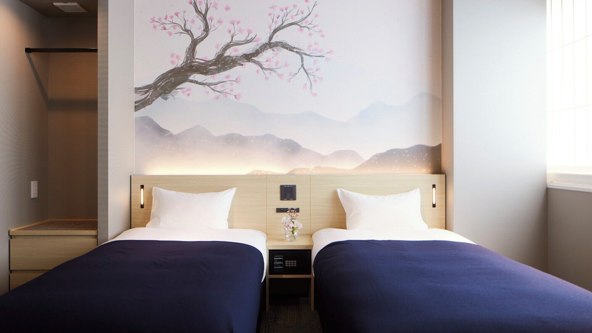 Habitación de hotel con dos camas, una mesita de noche, un panel de control en la pared y un cuadro decorativo de un árbol con flores rosadas.