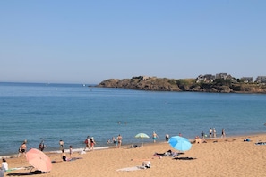 A beach with people sunbathing and swimming, a rocky coastline, and houses in the background.