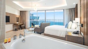 Habitación de hotel moderna con una cama grande, una zona de estar con una silla y una mesa, una bañera y un balcón con vistas a la ciudad.