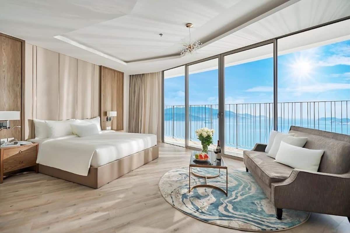 Habitación de hotel con una cama grande, un sofá, una mesita con un jarrón y fruta, y vista al mar a través de amplias ventanas.