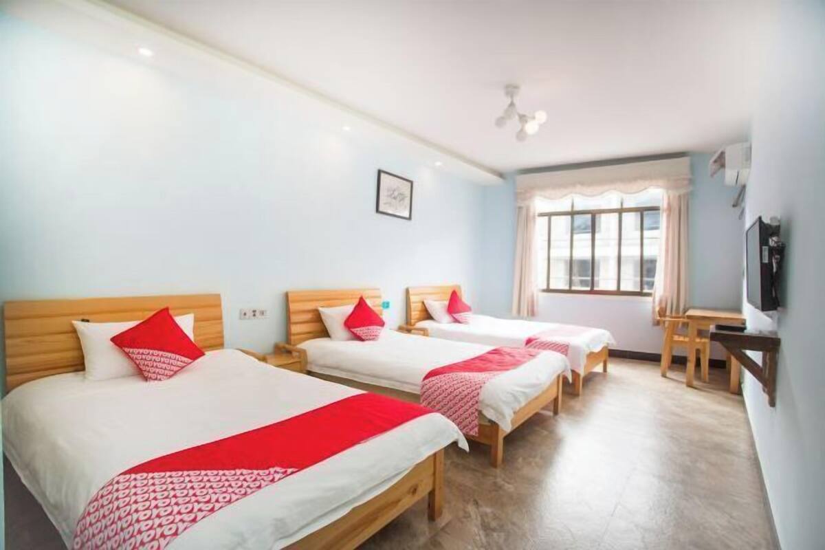 Une chambre d’hôtel avec trois lits simples, une tête de lit en bois, une télévision et une fenêtre avec des rideaux.