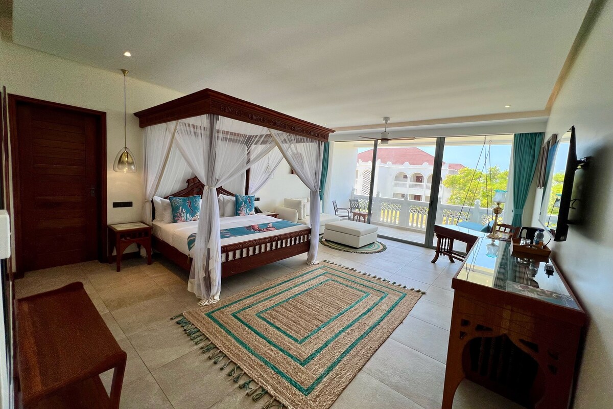 A spacious bedroom with a four-poster bed, a sitting area with a table and chairs, and a balcony with a view of a building and trees.