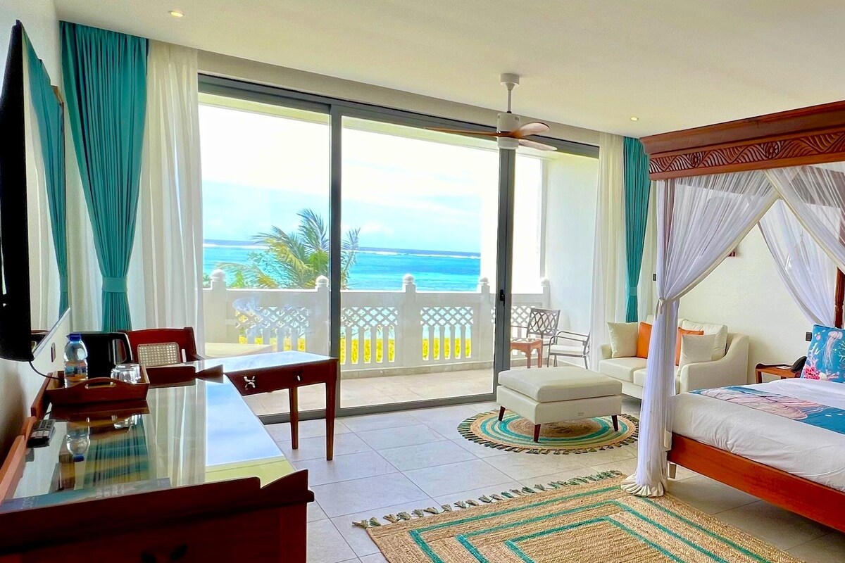 A bedroom with a four-poster bed, a balcony with a view of the ocean, and a dining area with a table and chairs.