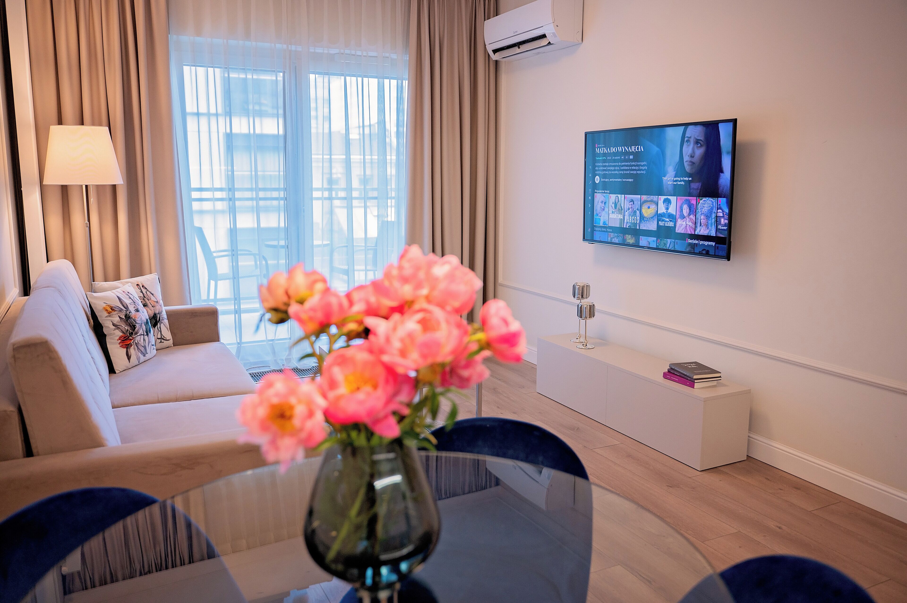Moderni hotellihuone, jossa on sohva, lasinen pöytä maljakko täynnä kukkia, seinälle asennettu taulutelevisio ja pieni valkoinen sivupöytä.