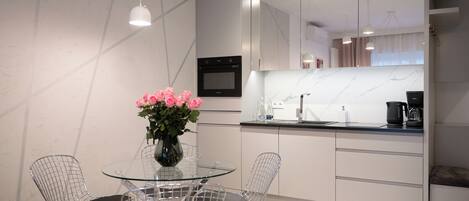 A modern kitchen with a marble countertop, stainless steel appliances, and a glass dining table with wire chairs.