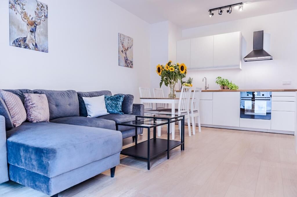 Una sala moderna con un sofá gris, una mesa de centro de vidrio y un comedor con sillas blancas y un jarrón de girasoles.