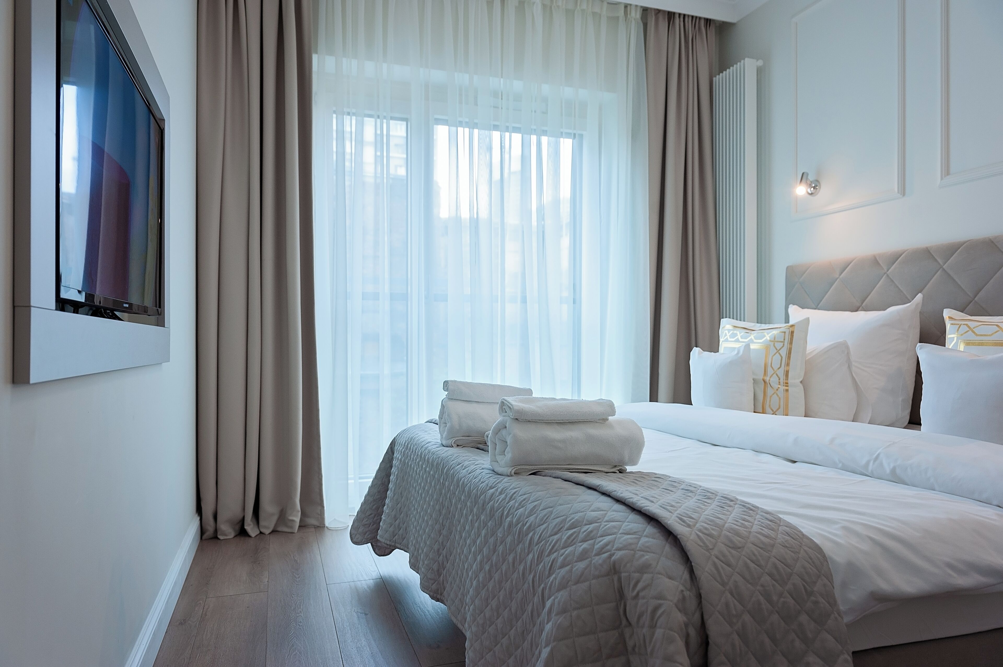 A neatly made bed with white linens and a quilted blanket, a large window with sheer curtains, and a wall-mounted light fixture.