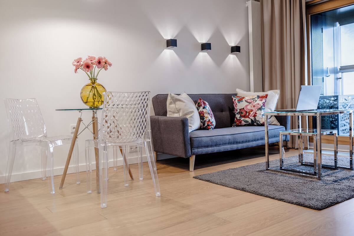 A modern living room with a grey sofa, clear chairs, a glass table, and a vase of flowers.