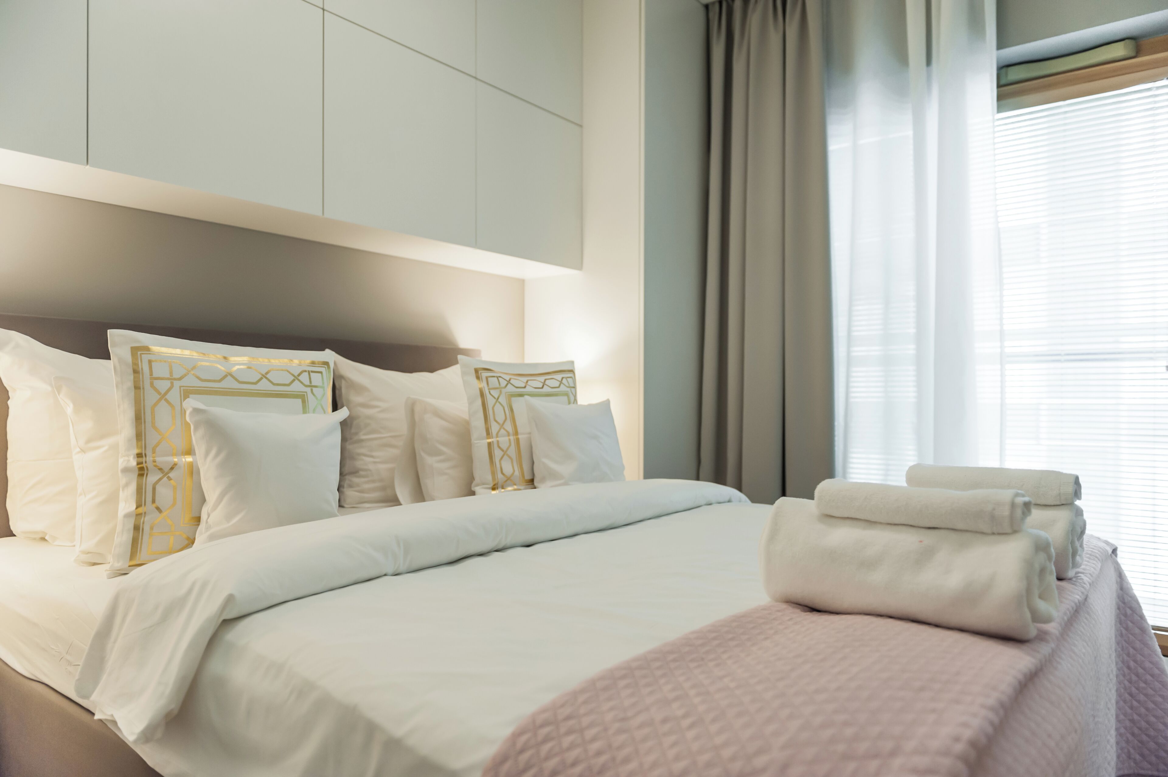 A neatly made bed with white bedding and decorative pillows, a pink blanket, and folded towels on the side.
