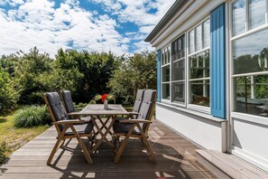 An outdoor wooden deck with a table and four chairs, a blue window, and a garden with trees and shrubs.