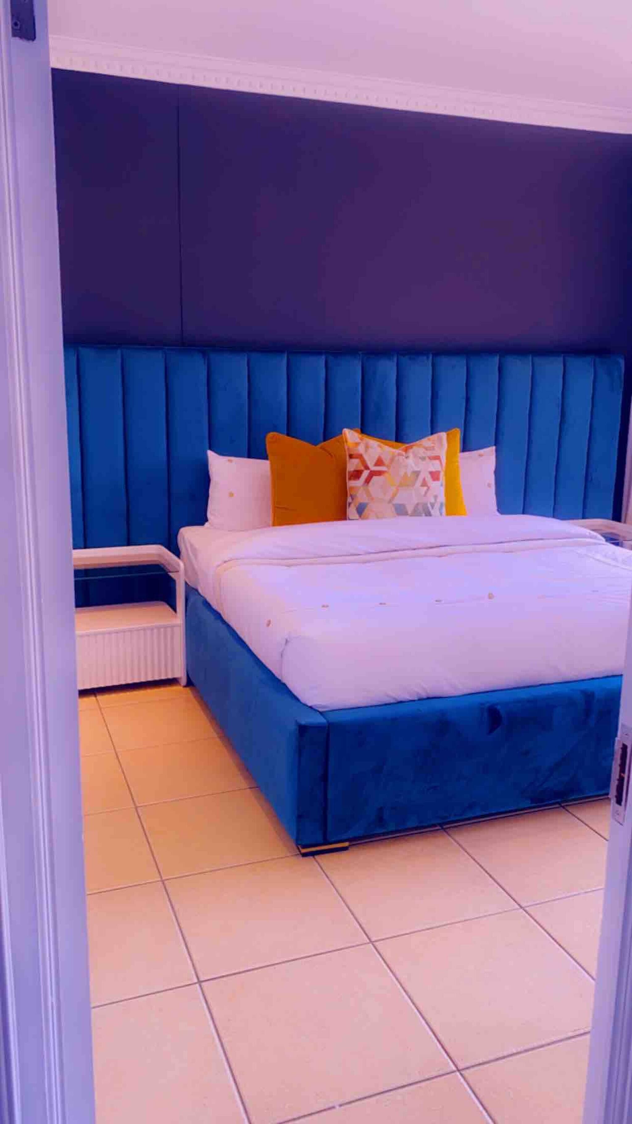 Un dormitorio con cabecera azul, cama con ropa de cama blanca y almohada colorida.
