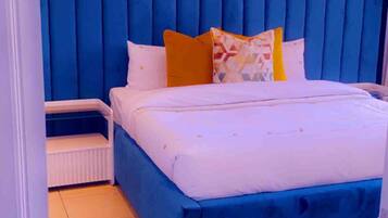 Un dormitorio con cabecera azul, cama con ropa de cama blanca y almohada colorida.