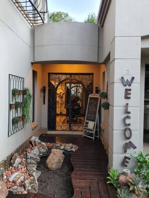 Fachada moderna de un edificio con un letrero que dice 'WELCOME', una reja metálica decorativa y un pequeño jardín con rocas y plantas.
