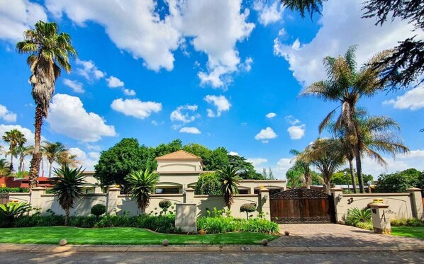 A large, single-story house with a prominent front gate, surrounded by lush greenery and palm trees. - Or Tambo Airport Holiday Home (Boksburg)