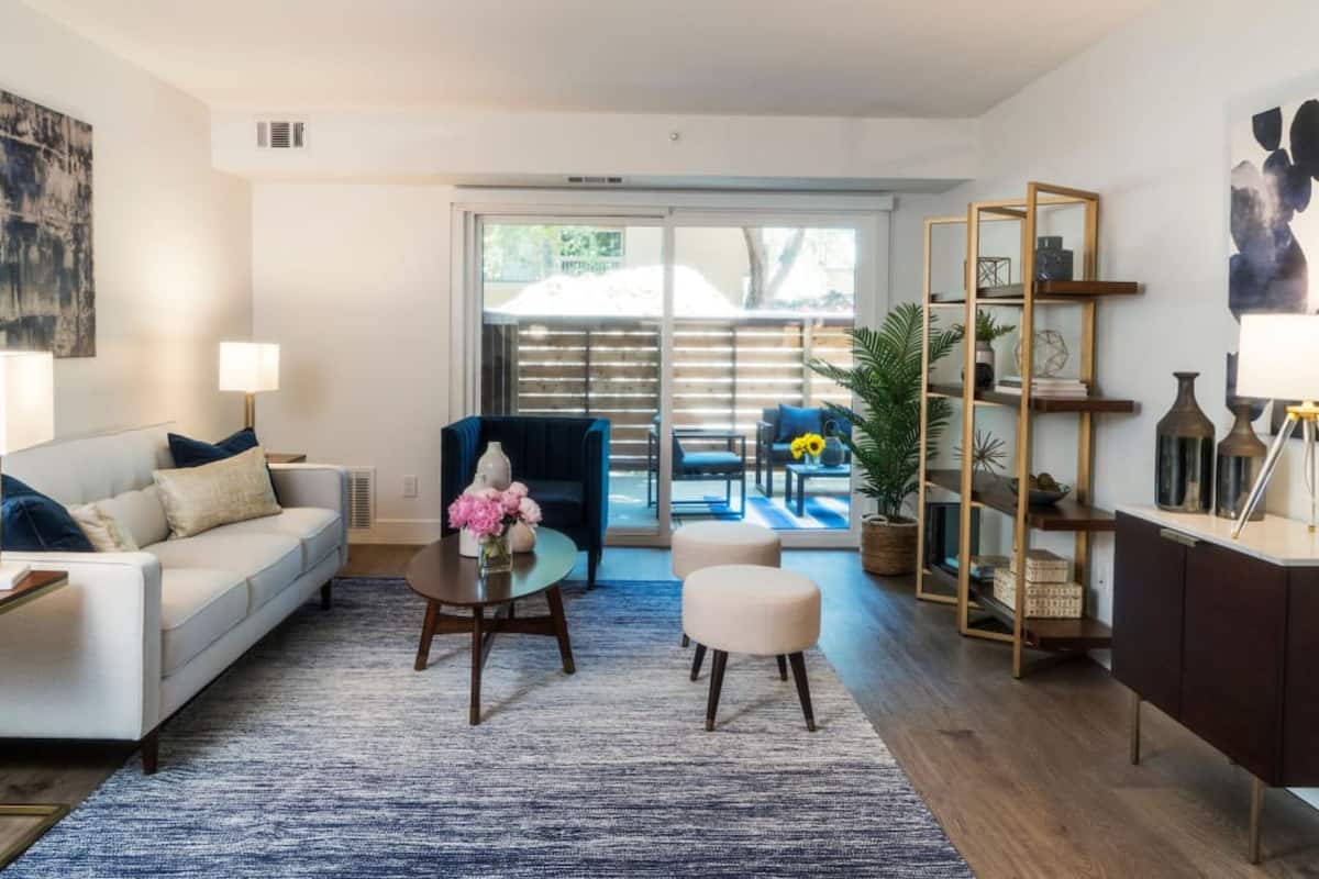 A modern living room with a sofa, coffee table, and ottoman, a glass door leading to a balcony, and a bookshelf.