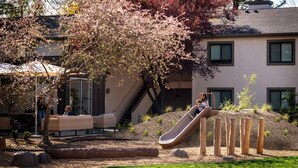 Un parque infantil con tobogán y estructuras para escalar, rodeado por un edificio con escaleras y una zona de estar cubierta.
