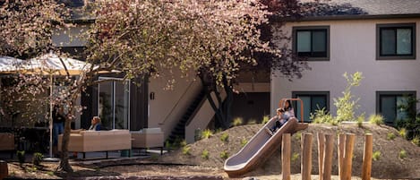 A playground with a slide and climbing structures, surrounded by a building with a staircase and a covered seating area.