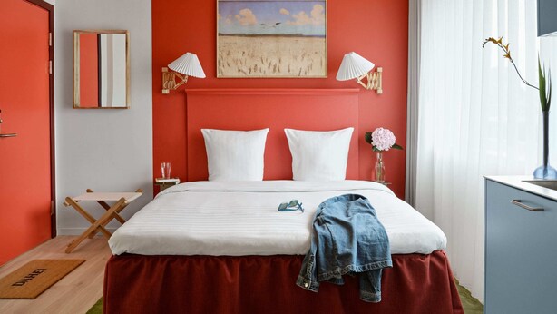A bedroom with a red accent wall, a bed with white linens, two bedside lamps, a painting above the bed, and a bedside table with a glass and books. - Cozy Studio Apartment for 2 (København)