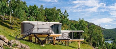 A modern RV with a flat roof and a covered deck area, situated on a grassy hillside with a lake and forested mountains in the background.