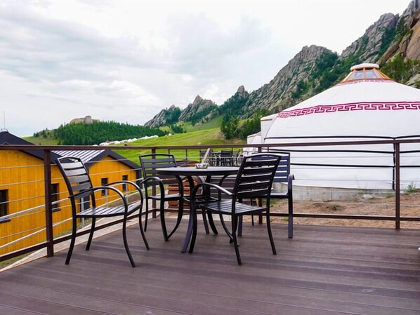 A wooden deck with a table and chairs, a yurt with a decorative pattern, and a mountain backdrop.