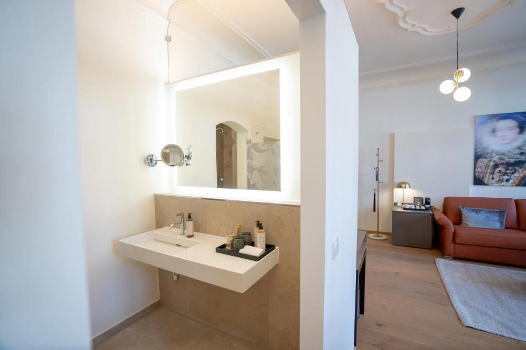 Un baño moderno con un espejo grande, un lavamanos blanco y un perchero. El suelo es de madera y en el área de estar contigua hay un sofá rojo.
