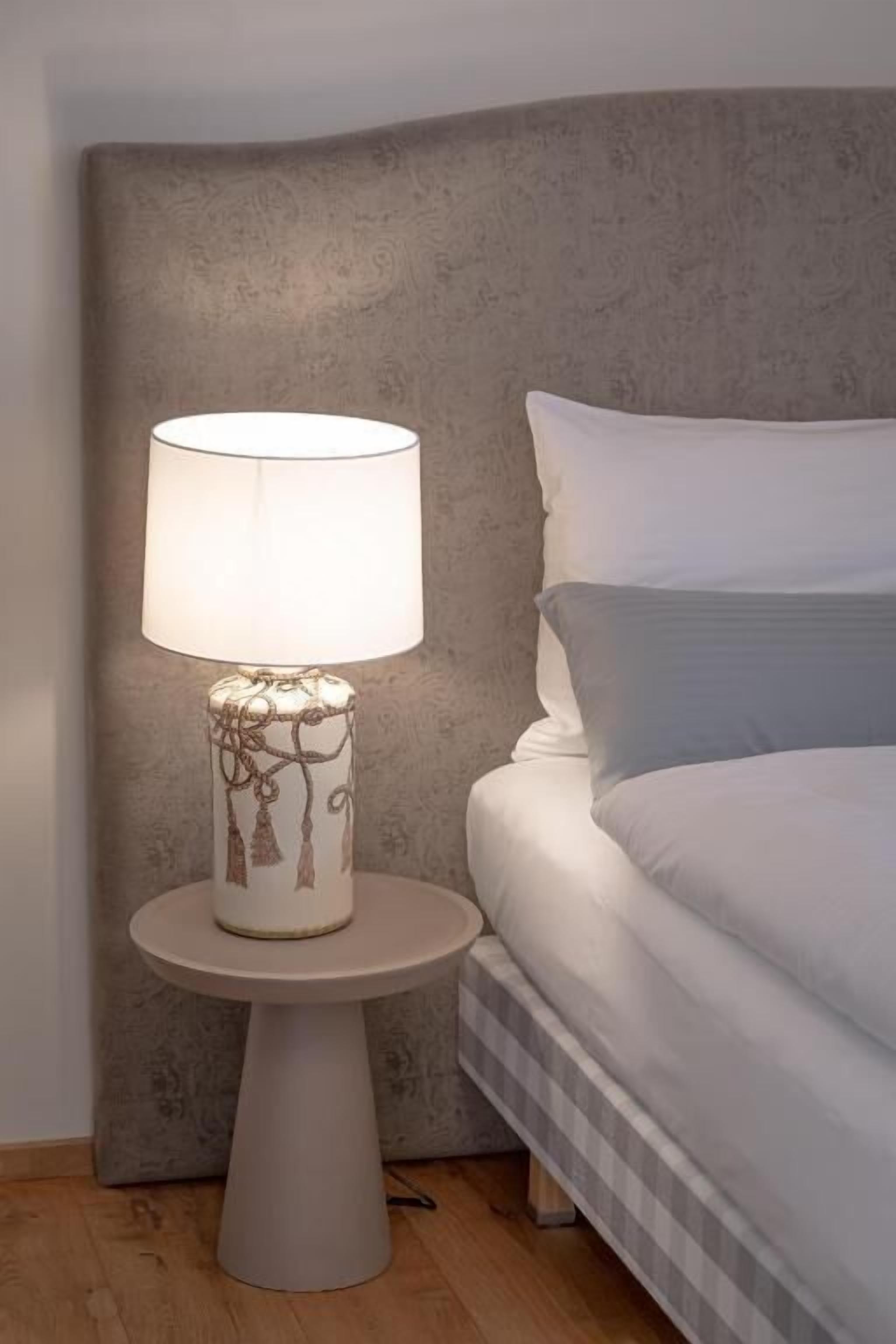 Una cama con ropa blanca y cabecera gris, una mesa redonda con una lámpara blanca con diseño de árbol, y suelo de madera.