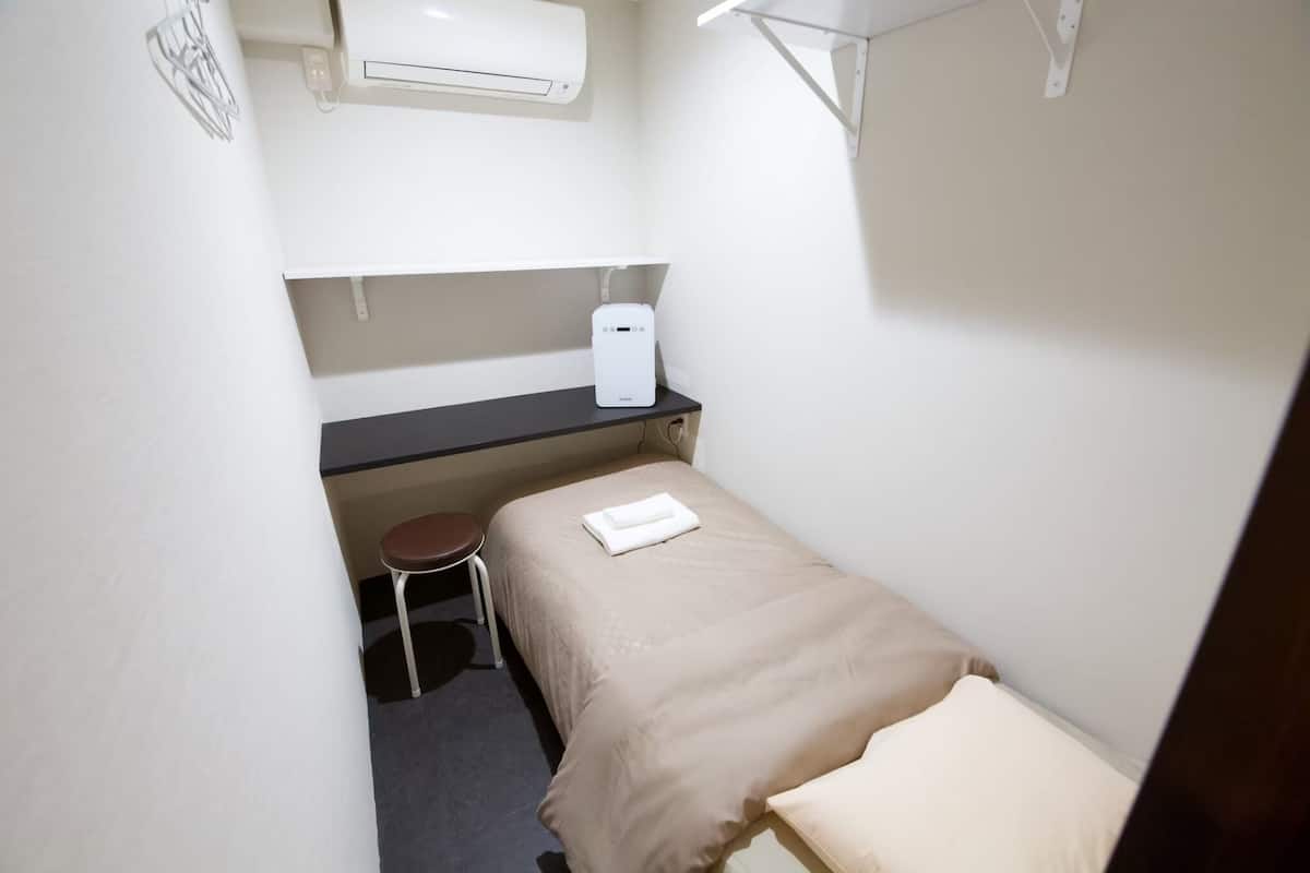 Una habitación pequeña y funcional con una cama individual, una mesita, una silla y un calentador de pared.