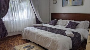 A neatly made bed with a white and purple bedspread, a fluffy rug with slippers, and a wooden floor. - Bliss Haven (Nairobi)