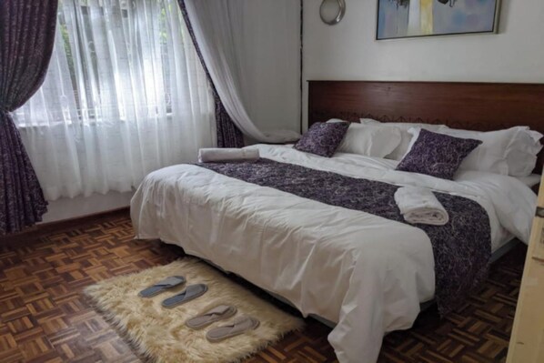 A neatly made bed with a white and purple bedspread, a fluffy rug with slippers, and a wooden floor. - Bliss Haven (Nairobi)