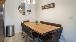 Un'area pranzo con un tavolo in legno, quattro sedie in rete metallica, una piccola pianta in vaso, uno specchio rotondo e un cartello con la scritta 'welcome' sulla parete.