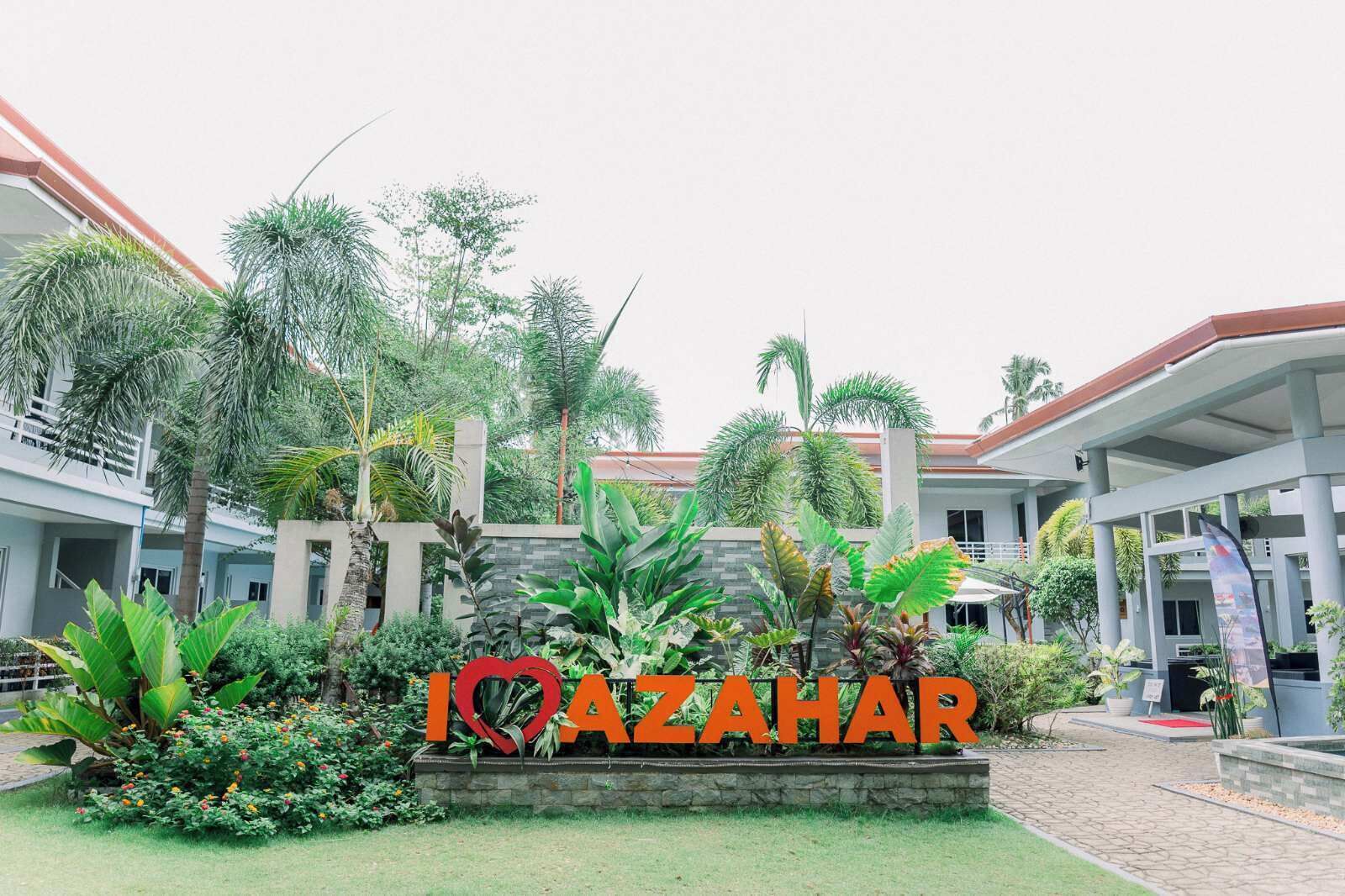 A courtyard with a sign reading "IOAZAHAR" surrounded by lush greenery and modern buildings.