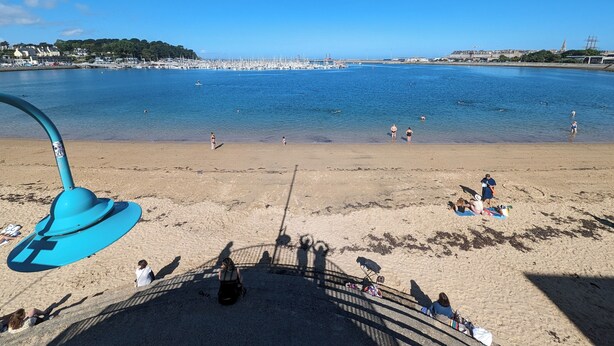 A beach with clear blue water, people swimming and sunbathing, and a distant view of a marina with boats.