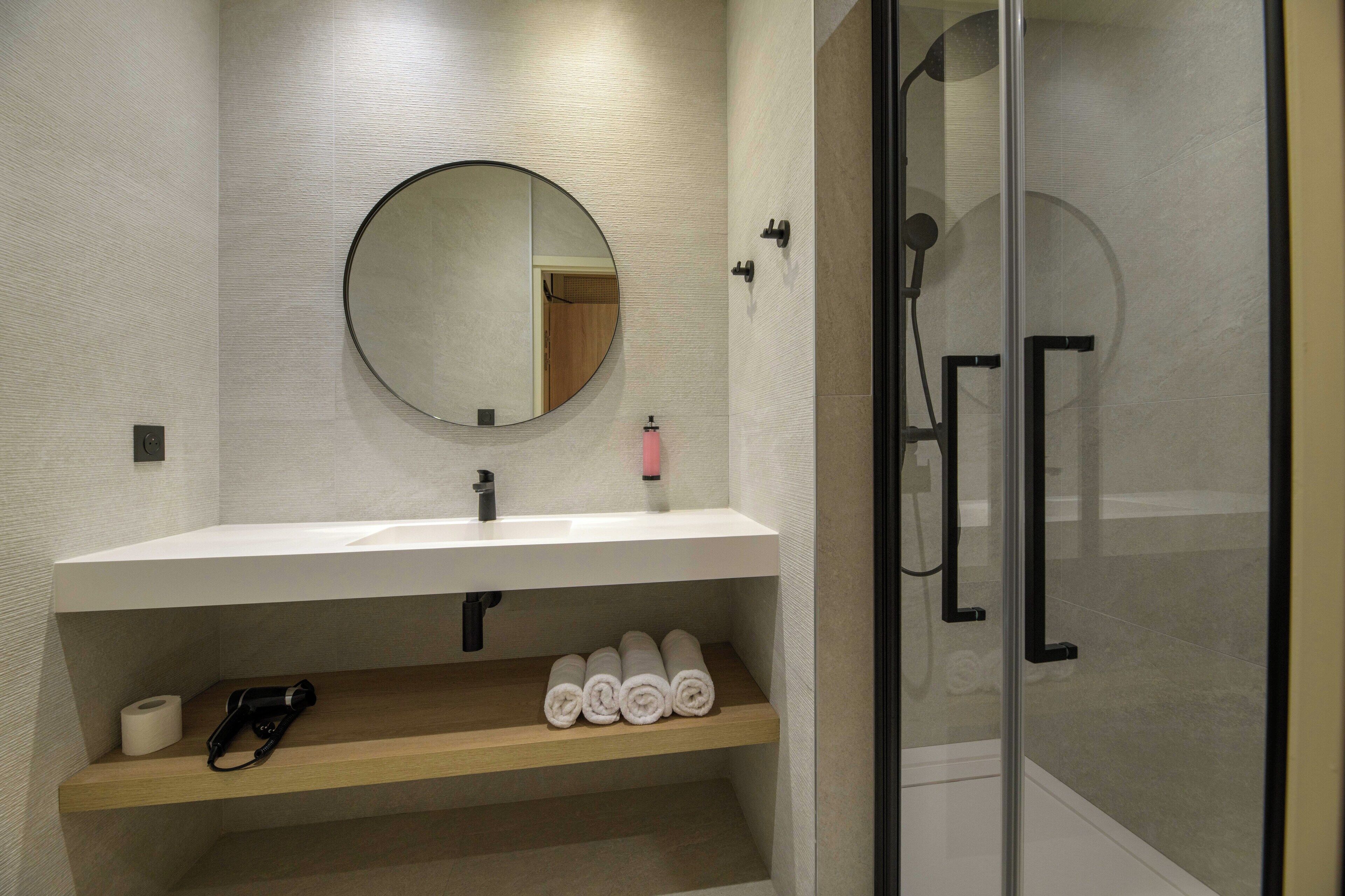 Un baño moderno con un lavamanos blanco, un espejo redondo, una mampara de vidrio para la ducha y toallas ordenadamente dobladas en un estante.