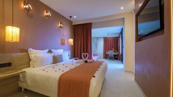 A hotel room with a bed, bedside tables, a TV, and a view into the dining area.