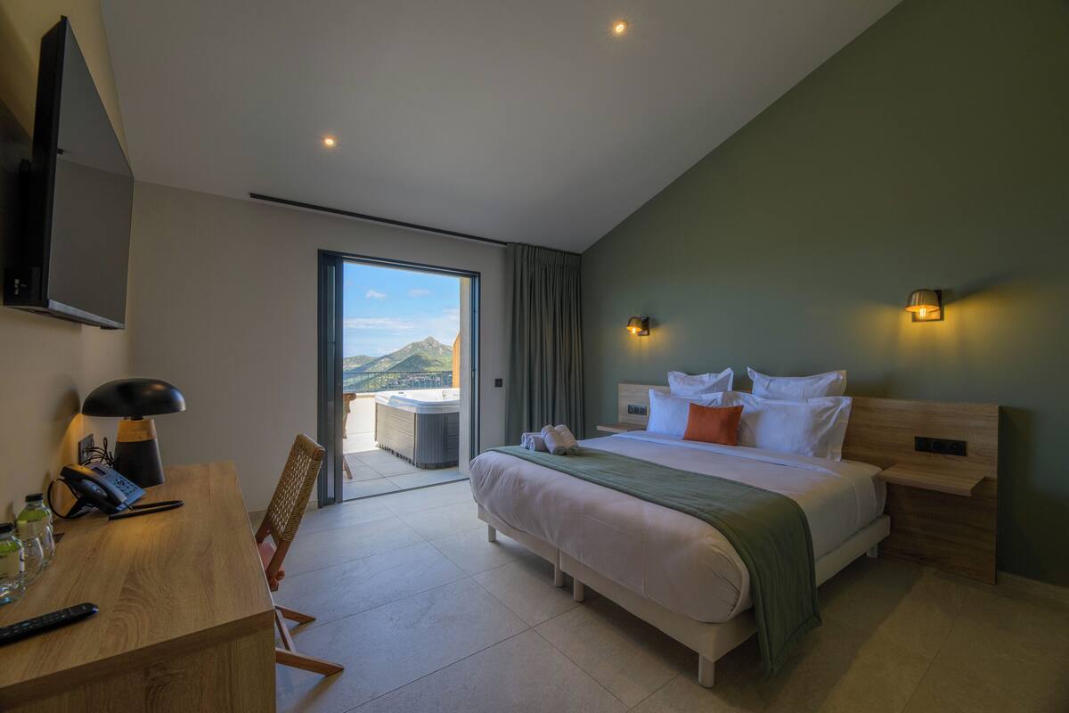 A modern hotel room with a large bed, a desk with a lamp, and a view of the mountains through the window.