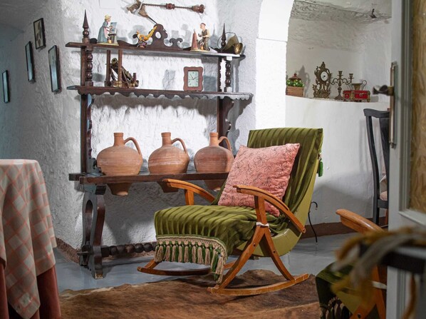 A rustic room with a wooden rocking chair, a shelf with pottery, and a wall clock. - Cueva Navarro      . (Gorafe)