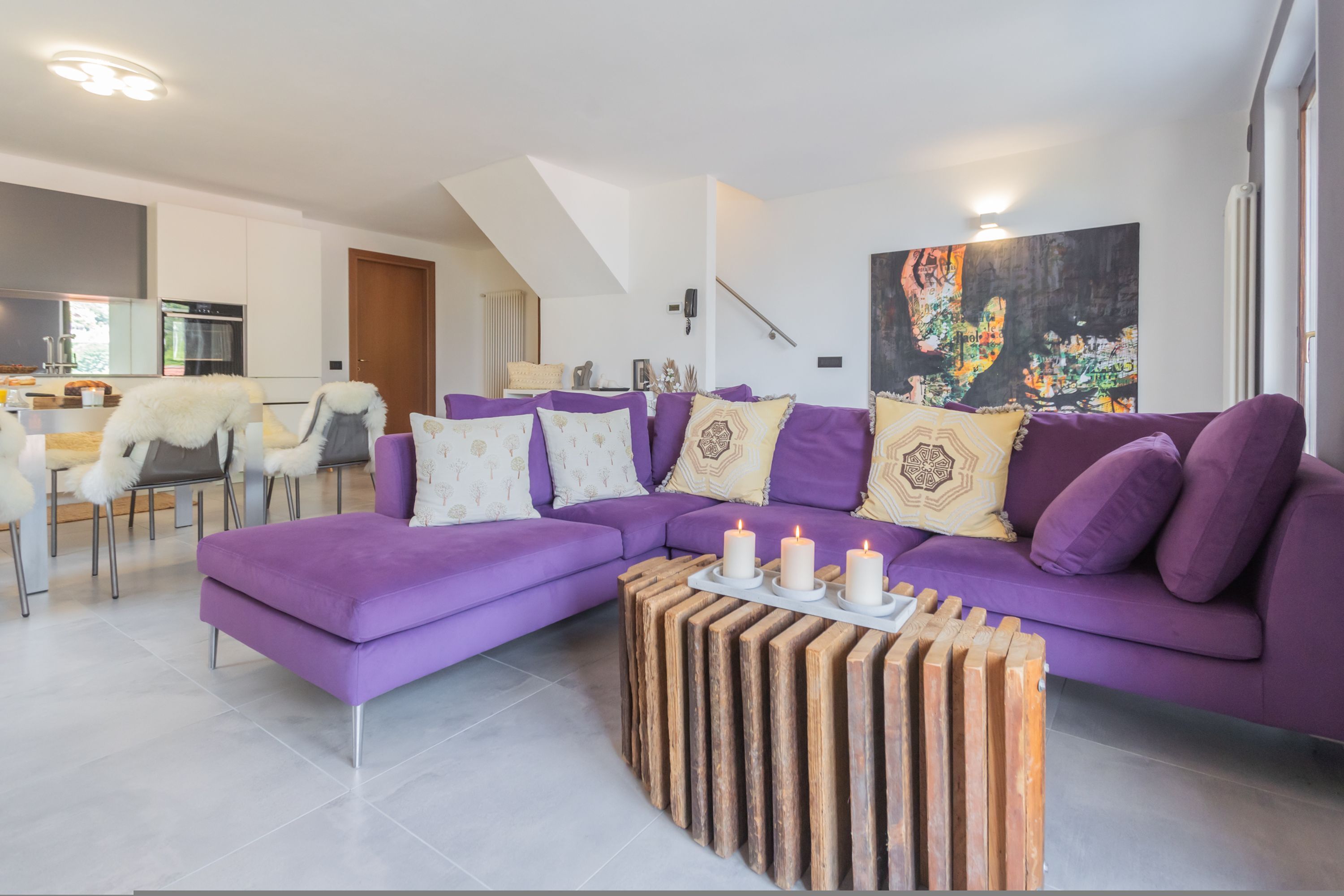 Un salon moderne avec un canapé d’angle violet, une table basse en bois et un grand tableau abstrait accroché au mur.