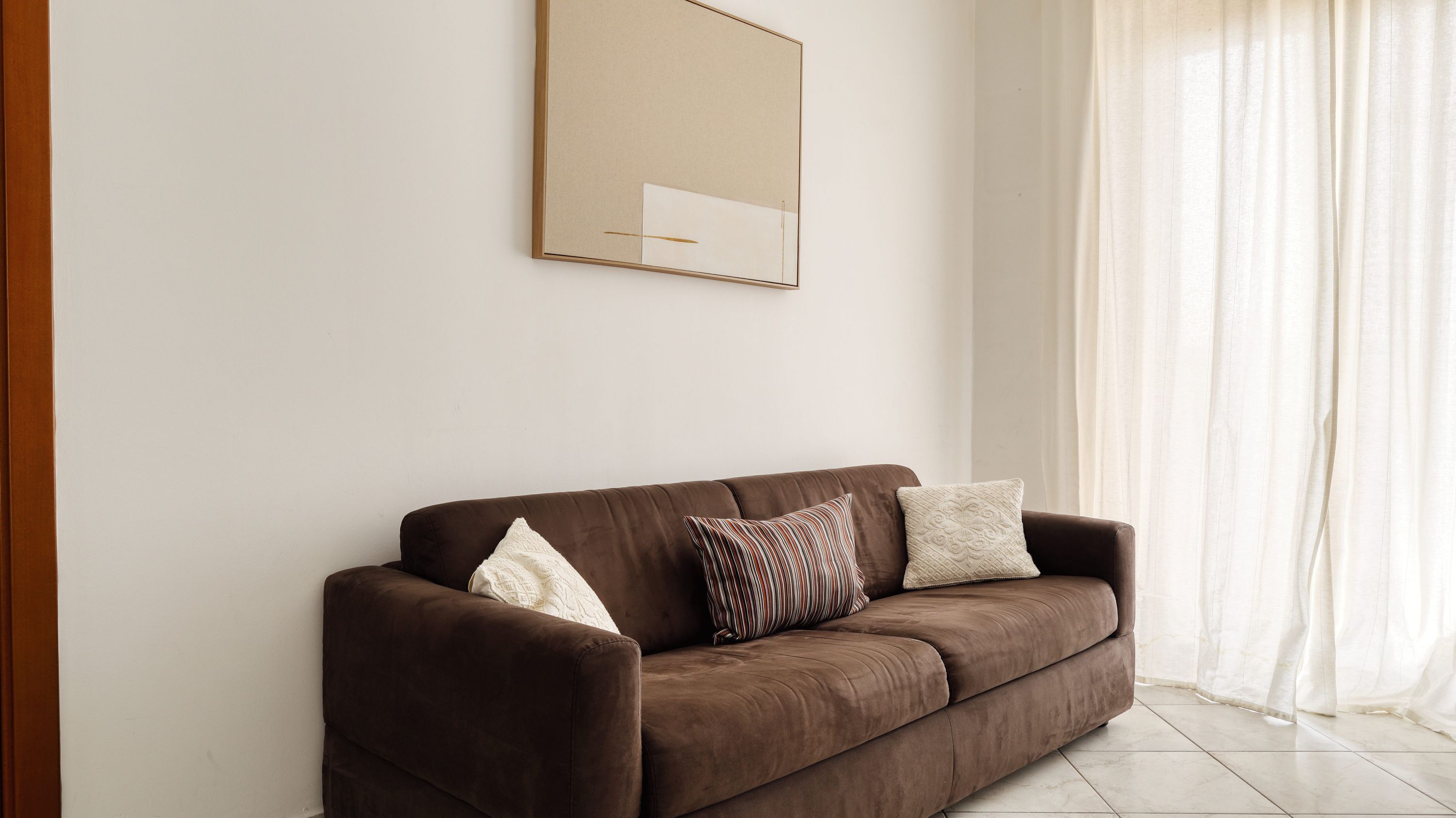 A brown sofa with two pillows against a plain wall, a framed picture above it, and sheer curtains on a window.