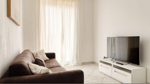 Ett vardagsrum med en brun soffa, vita gardiner och en platt-TV placerad på ett vitt skåp.