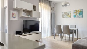 Ett modernt vardagsrum med en platt-TV, en matplats med bord och stolar samt ett stort fönster med tunna, genomskinliga gardiner.