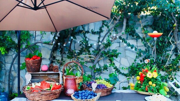 Une table sur laquelle on trouve différents paniers remplis de fruits et de légumes, une lampe décorative et un mur de pierre avec de la verdure en arrière-plan.