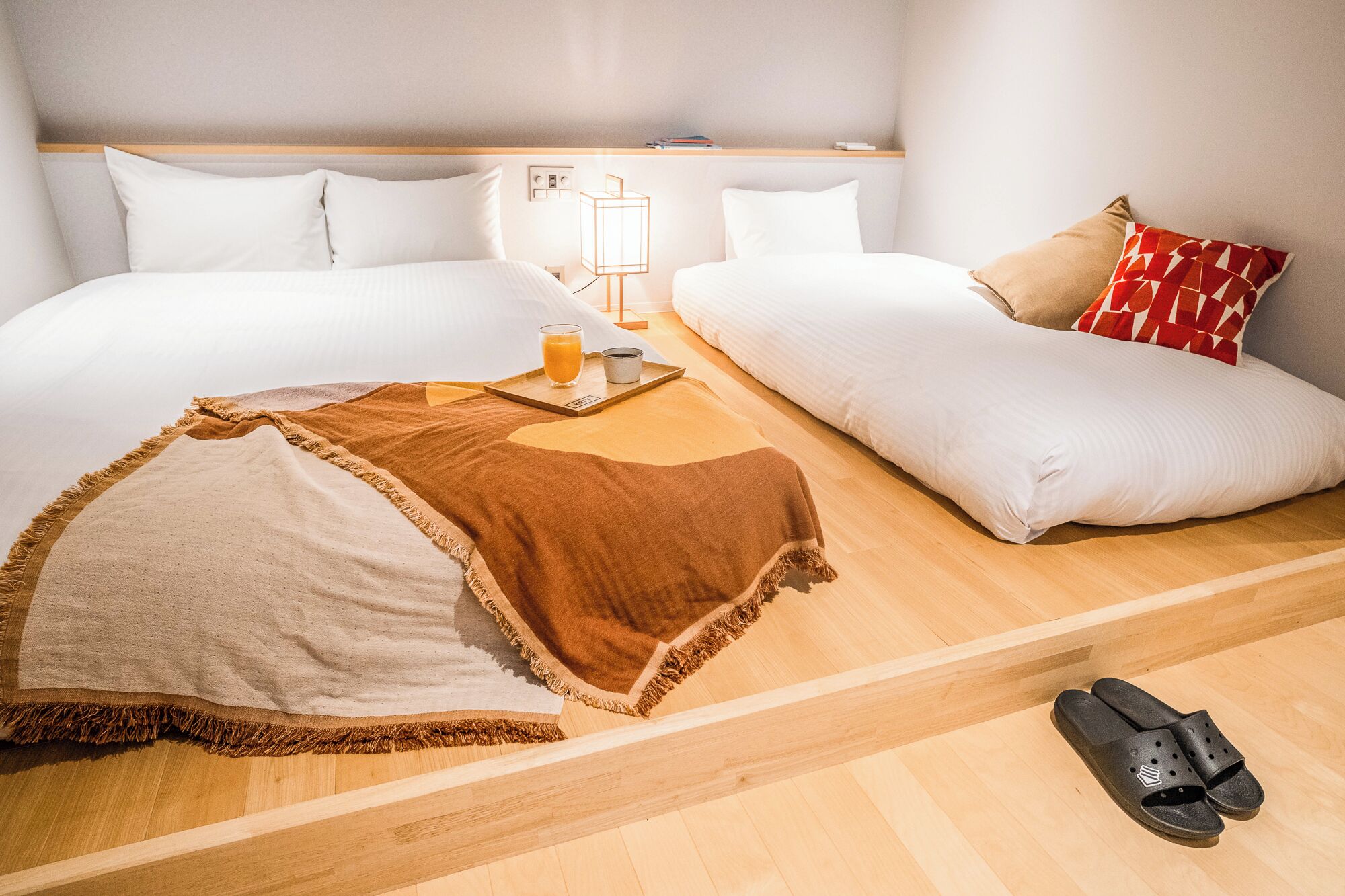 A hotel room with a bed, a sofa, a wooden floor, a blanket, a tray with a glass of orange juice, and a pair of black slippers.