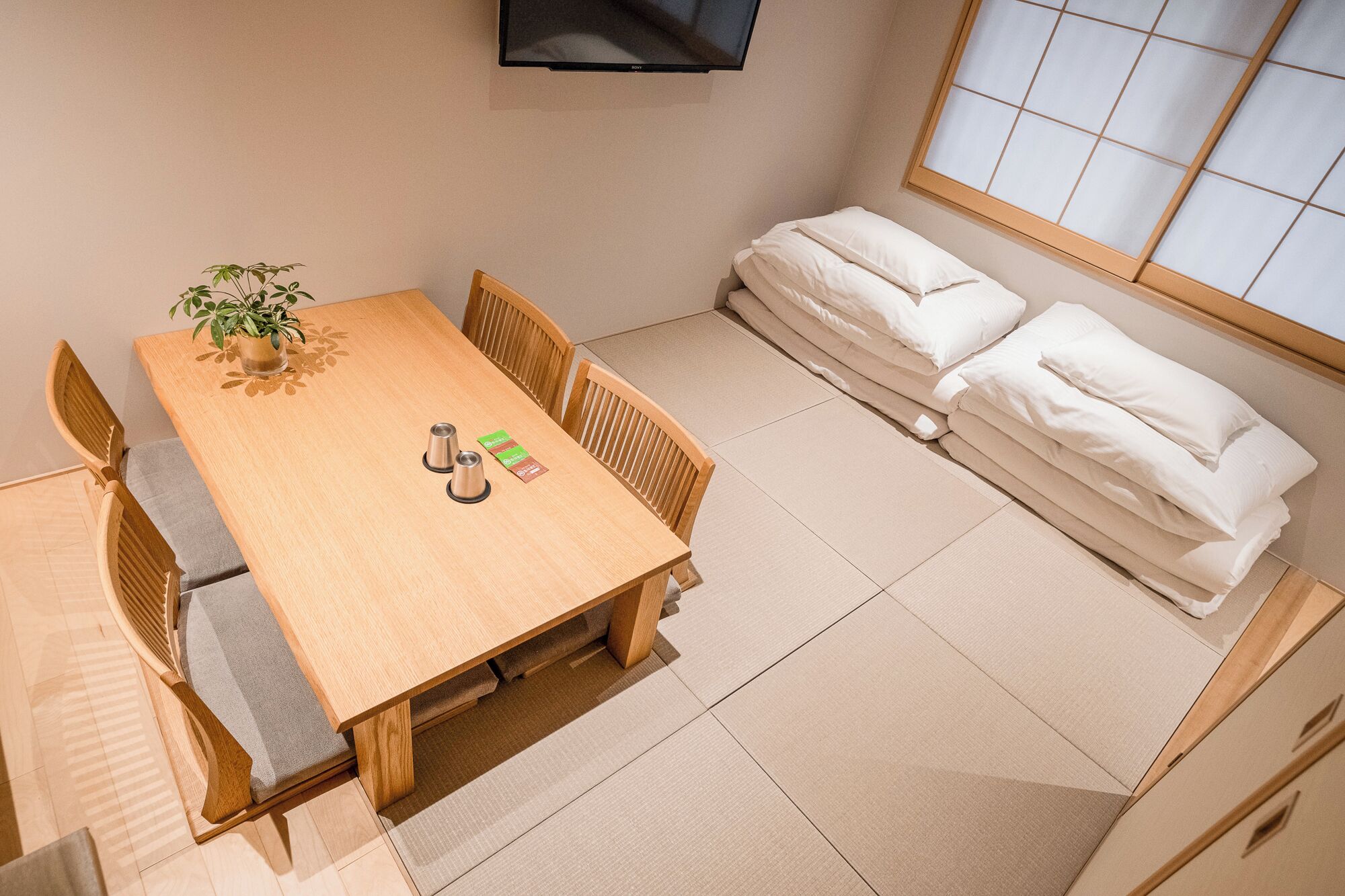 Une pièce de taille modeste, comprenant une table à manger en bois, des chaises, une petite plante en pot et un futon servant de lit, agrémenté de coussins.